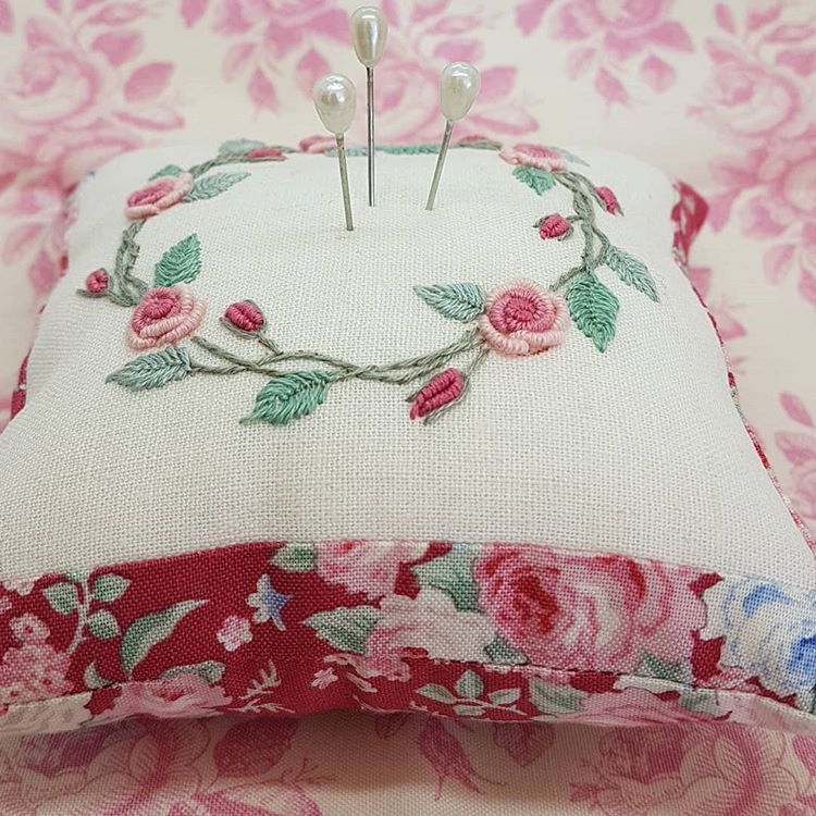 An embroidered linen pin cushion with pink floral embroidery on top and a red floral fabric trim, holding three pins, set against a matching pink floral background.
Photo by Angela Watson [additional_image_link]