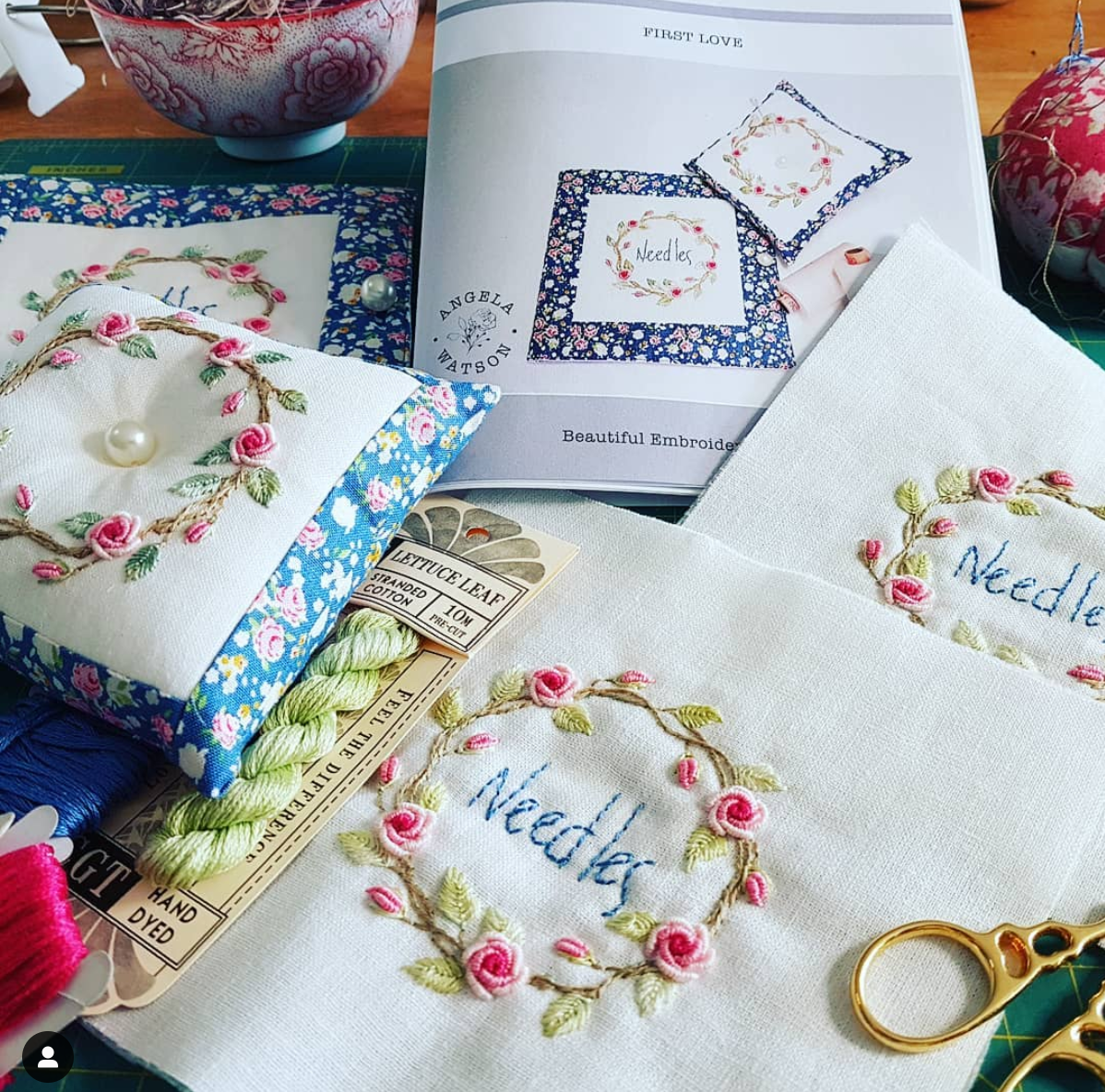 A close-up of embroidered fabric pieces and sewing supplies on a table, including white fabric with pink rose floral borders and the word 'Needle' stitched in blue, a small embroidered pillow, spools of thread, scissors, and an open embroidery book.
Photo by Angela Watson [additional_image_link]