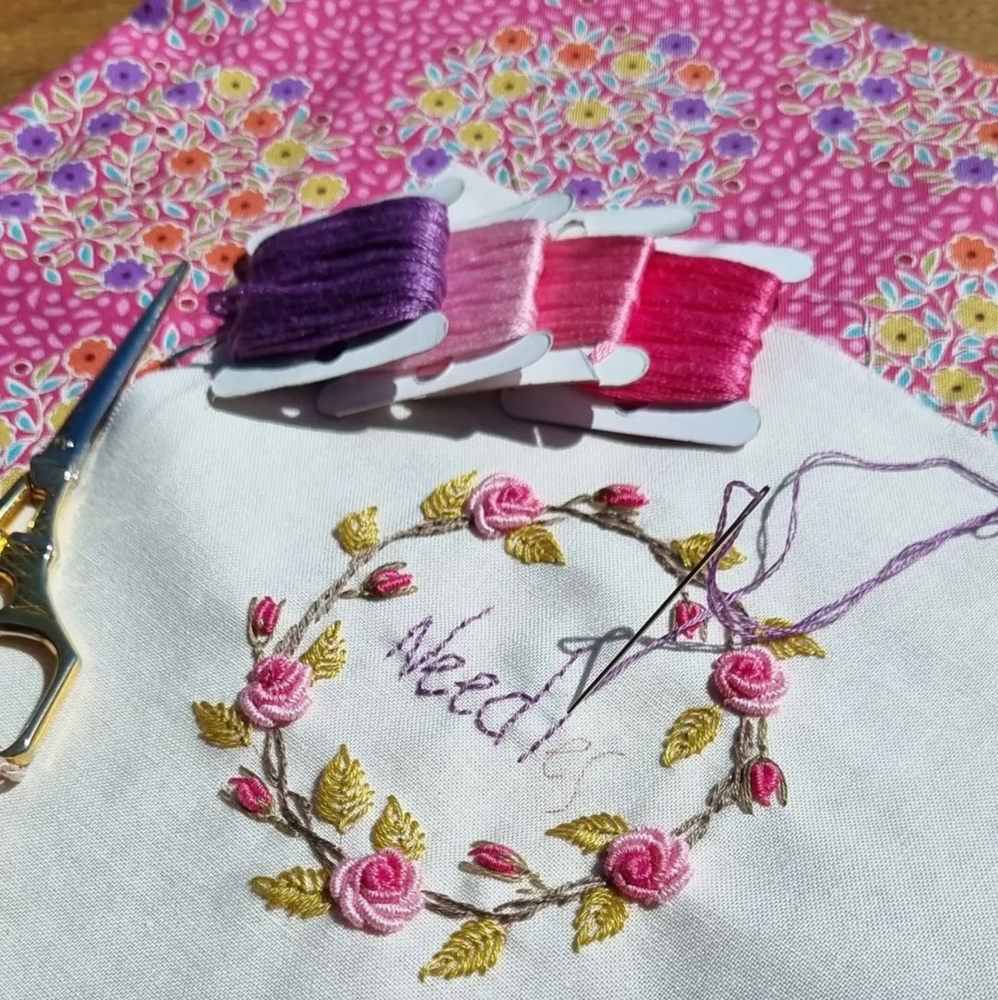 A close-up of a handmade embroidered needle with pink and gold floral thread, placed on a white fabric surface next to scissors and three spools of embroidery floss in purple, pink, and red. A pink patterned fabric background is visible.
Photo by Angela Watson [additional_image_link]