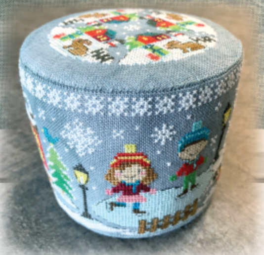 A round, low-profile pouf ottoman featuring a light blue winter-themed cross-stitch pattern with children ice skating, snowflakes, and a festive top design.
Photo by Tiny Modernist [additional_image_link]