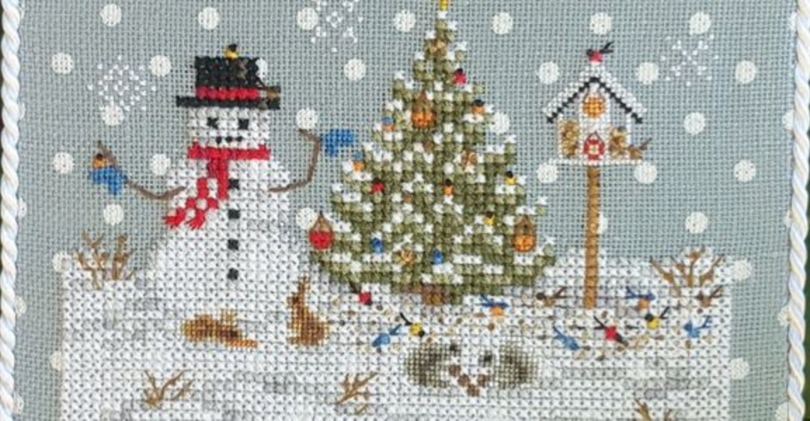 A cross stitch kit pattern depicting a Christmas ornament with a snowman, a Christmas tree, and decorative elements on a blue linen background with white polka dots.
Close up.
Photo by Blackberry Lane Designs [additional_image_link]