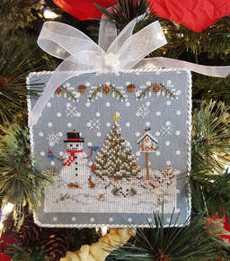 A cross stitch kit pattern depicting a Christmas ornament with a snowman, a Christmas tree, and decorative elements on a blue linen background with white polka dots.
Photo by Blackberry Lane Designs [additional_image_link]