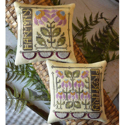 Two square embroidered decorative pillows displayed on woven baskets with green fern leaves. The top pillow has a floral design with the word 'FARMLAND' on the left, and the bottom pillow has a multi-flower design with the word 'FARMLAND' on the right.
[image_link]