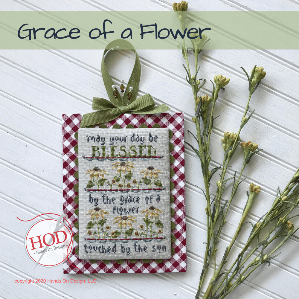 A decorative hanging ornament with a red-and-white gingham border, a green ribbon bow, and embroidered text reading 'May your day be BLESSED by the grace of a flower touched by the sun,' shown on a white paneled background with dried flowers and a 'Grace of a Flower' title banner.
[image_link]