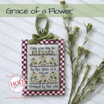 A decorative hanging ornament with a red-and-white gingham border, a green ribbon bow, and embroidered text reading 'May your day be BLESSED by the grace of a flower touched by the sun,' shown on a white paneled background with dried flowers and a 'Grace of a Flower' title banner.
[image_link]