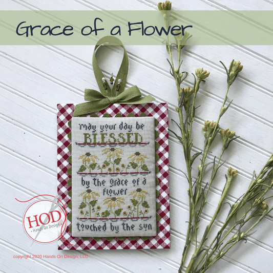 A decorative hanging ornament with a red-and-white gingham border, a green ribbon bow, and embroidered text reading 'May your day be BLESSED by the grace of a flower touched by the sun,' shown on a white paneled background with dried flowers and a 'Grace of a Flower' title banner.
[image_link]