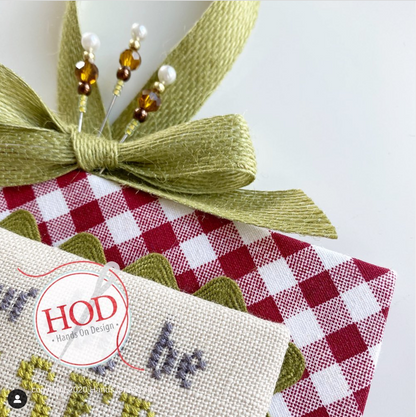 A close-up of a decorative gift box wrapped in red and white gingham fabric, tied with an olive green ribbon and adorned with three beaded pins. A portion of a cream-colored cross-stitch piece is visible in the foreground with a circular 'HOD Hand On Design' watermark.
[additional_image_link]