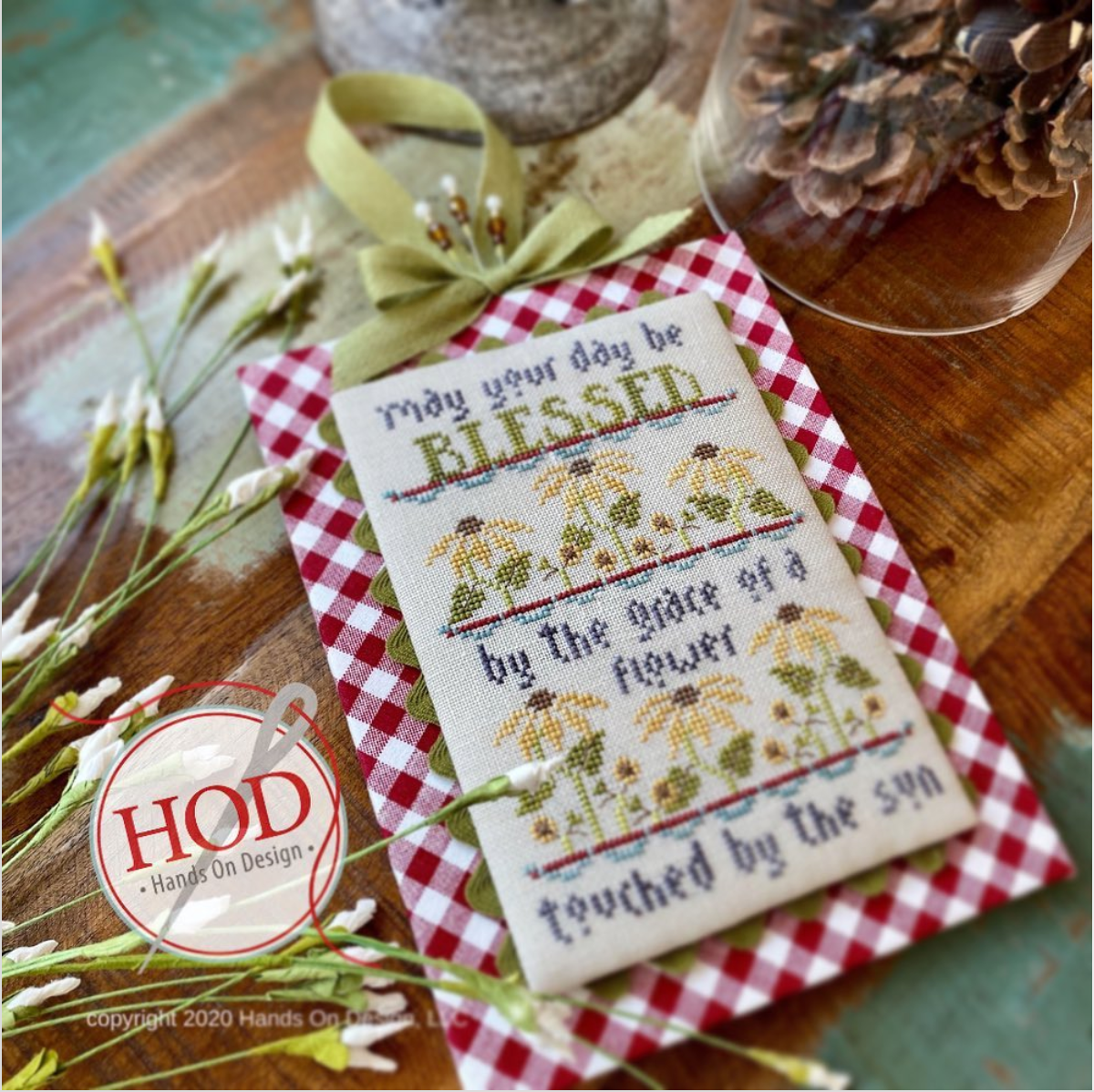 A framed cross-stitch piece with the words 'May your day be BLESSED by the grace of a flower touched by the sun' and floral embroidery, styled on a wooden surface with a red-and-white gingham border, green ribbon, pinecones, and small flowers. A circular 'HOD - Hands On Design' watermark appears in the lower left.
[additional_image_link]