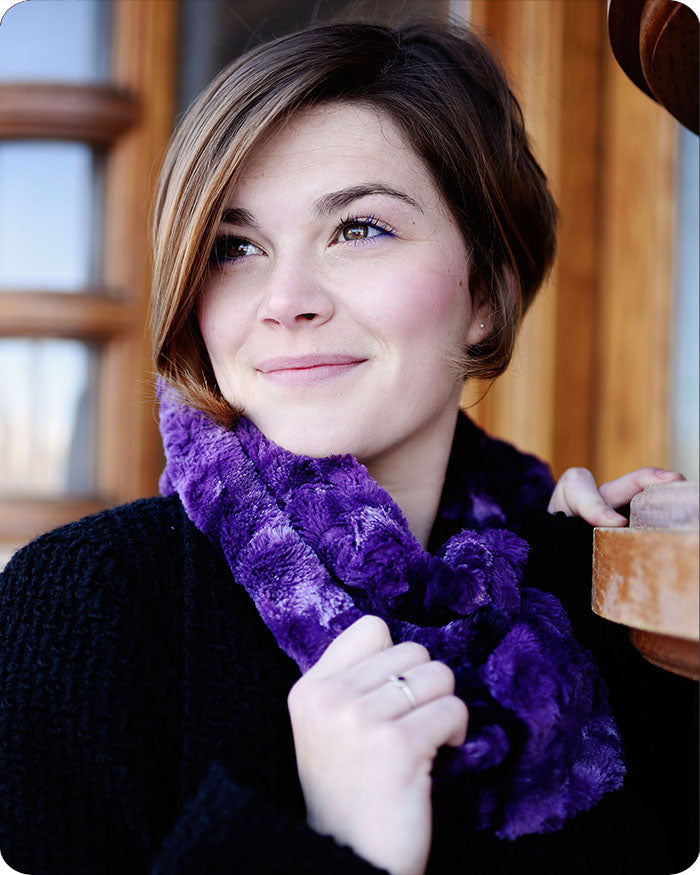 A woman with short brown hair wearing a plush, textured purple scarf and a black knit sweater, smiling while holding a wooden mug in a warm indoor setting.
Photo by Shannon Fabrics [additional_image_link]