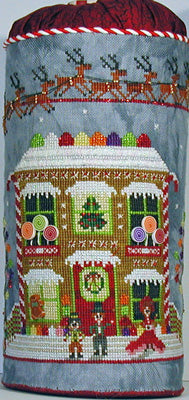 A cylindrical needlepoint canvas featuring a festive Christmas village scene with a gingerbread house, reindeer, and snow-covered figures on a gray-blue background.
Photo by PraiseworthyStitches.com [image_link]