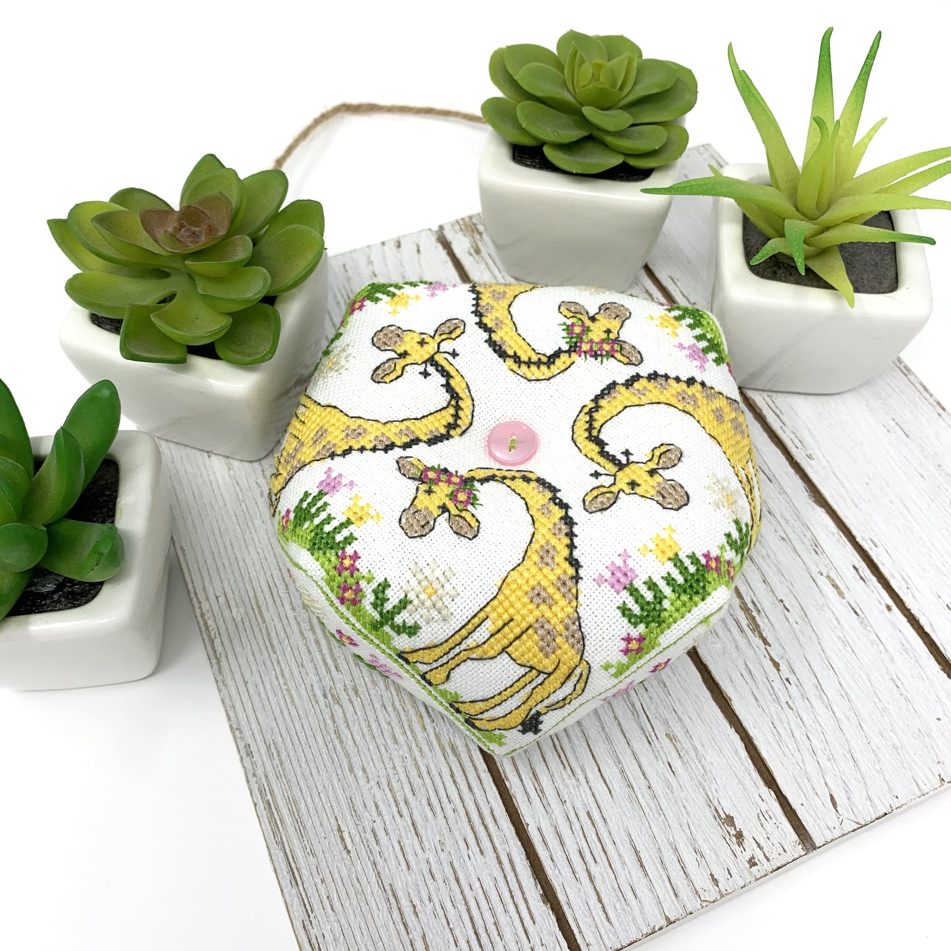 A hexagonal fabric-covered item, likely a small box or cushion, featuring a playful giraffe print on a white background. It is styled on a white wooden board surrounded by small potted green succulents.
Photo by Tiny Modernist [image_link]