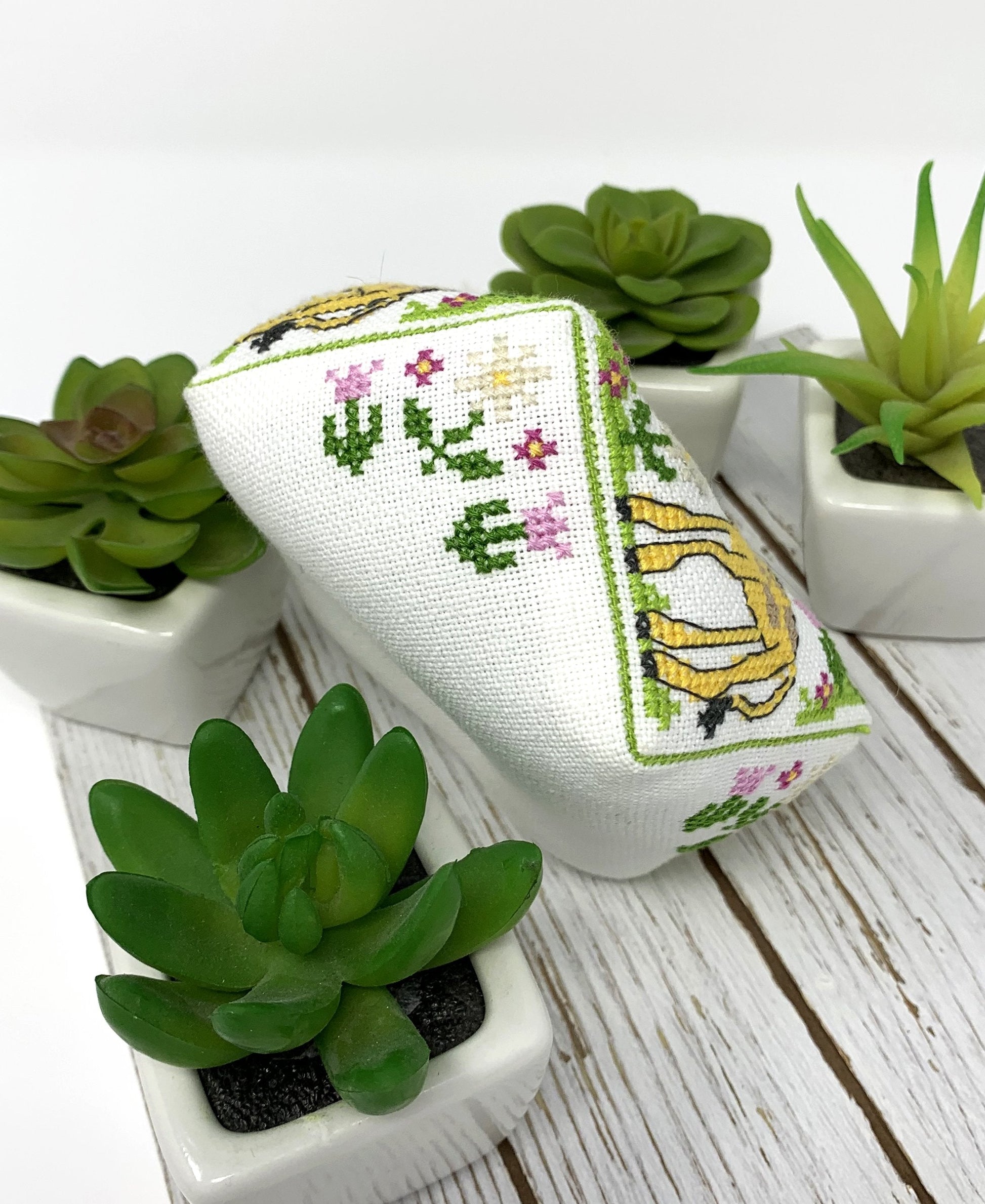 A small white embroidered fabric pouch or cushion with a giraffe and floral cross-stitch design, styled on a white wood surface with small potted succulents around it.
Photo by Tiny Modernist [additional_image_link]