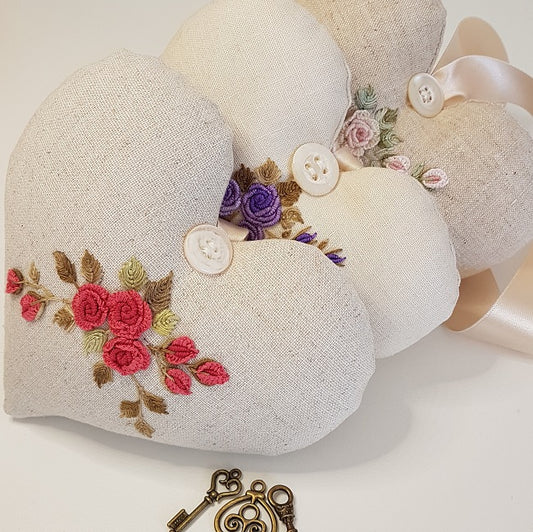 Three round linen heart-shaped pillow keepsakes with embroidered roses in pink, purple, and red, decorated with white buttons and ribbons, displayed on a white surface with antique keys.
Photo by Angela Watson [image_link]