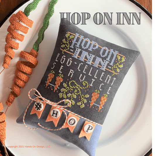 Decorative pillow with 'Hop on Inn' text and cross-stitched design, surrounded by decorative carrots on a white plate.
Photo by HandsOnDesign.biz [image_link]