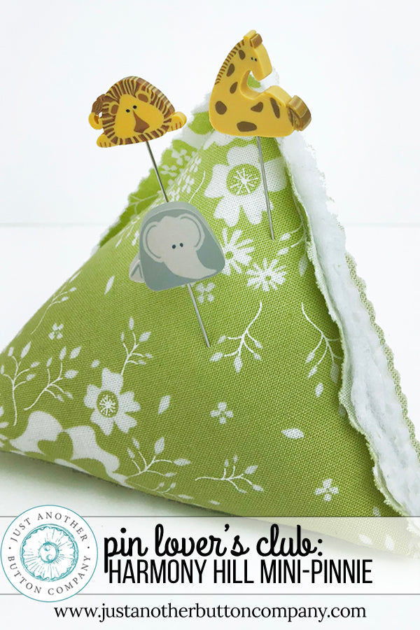A handmade fabric pin holder shaped like a mini-pinnie, featuring a green and white floral print on the exterior and a soft white fleece lining. It is styled with three decorative animal-shaped pins (lion, giraffe, elephant) and includes a text banner at the bottom with branding.
Photo by JustAnotherButtonCompany.com [image_link]