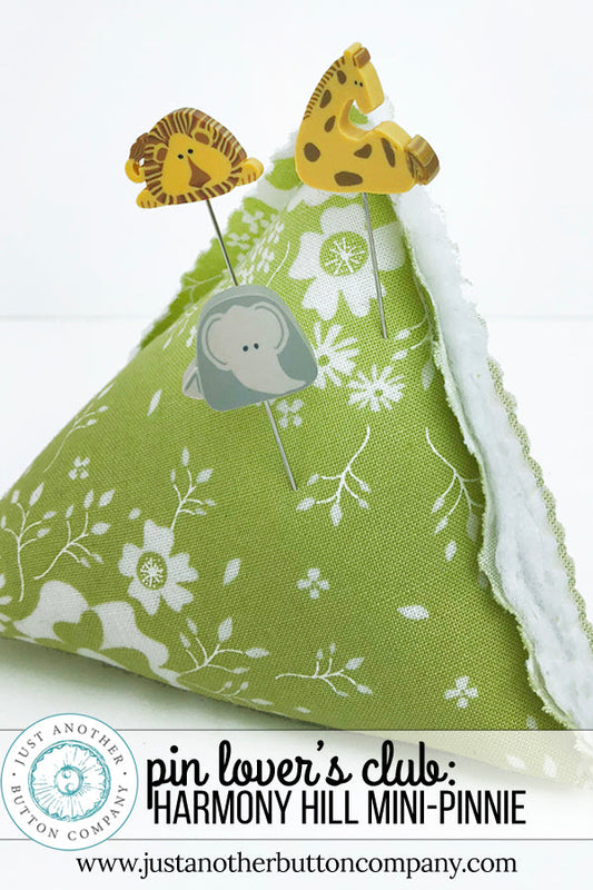 A handmade fabric pin holder shaped like a mini-pinnie, featuring a green and white floral print on the exterior and a soft white fleece lining. It is styled with three decorative animal-shaped pins (lion, giraffe, elephant) and includes a text banner at the bottom with branding.
Photo by JustAnotherButtonCompany.com [image_link]
