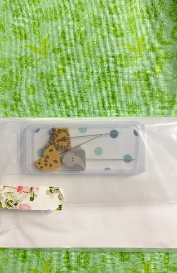 A small clear plastic case containing decorative pins or charms, placed on a white sheet over a green leaf-patterned fabric background. A floral washi tape strip is visible at the lower left.
Photo by TheSurgeonsKnots.com [additional_image_link]