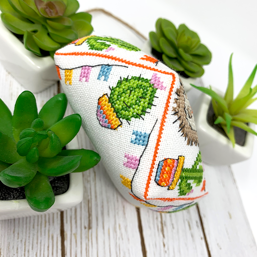A small cross-stitch cushion with a cactus and floral pattern, styled on a white wooden surface with green succulent plants in white pots.
Photo by Tiny Modernist [additional_image_link]