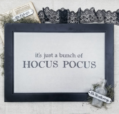 A framed decorative sign with the text 'it's just a bunch of HOCUS POCUS' in dark lettering on a light background. The frame is dark charcoal/black, and the sign is styled with black lace trim, a vintage bottle, moss, and paper props on a textured fabric surface.
Photo by Hellofromlizmathews.com [image_link]