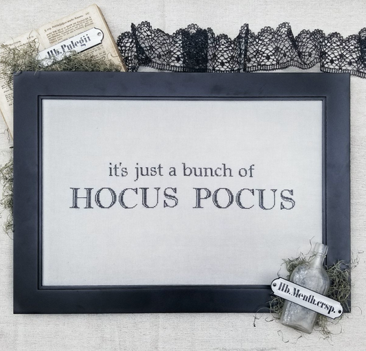 A framed decorative sign with the text 'it's just a bunch of HOCUS POCUS' in dark lettering on a light background. The frame is dark charcoal/black, and the sign is styled with black lace trim, a vintage bottle, moss, and paper props on a textured fabric surface.
Photo by Hellofromlizmathews.com [image_link]