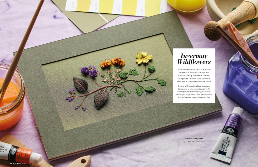 A framed botanical art piece featuring pressed wildflowers on a textured olive-green background, styled on a lavender surface with paint tubes, brushes, and art supplies around it. A text panel reads 'Invermary Wildflowers.
Photo by InspirationsStudios.com [additional_image_link]