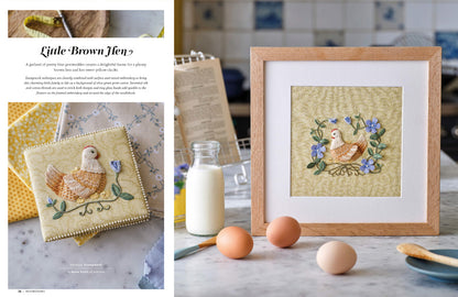 A framed handmade quilt featuring a brown hen and blue flowers on a beige background, displayed in a light wood frame on a kitchen counter next to eggs, a glass bottle of milk, and a cookbook. The left page of a book shows the title 'Little Brown Hen'.
Photo by InspirationsStudios.com [additional_image_link]