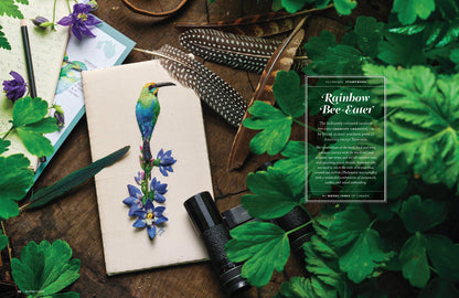 A styled flat-lay featuring a cream-colored sketchbook with a detailed illustration of a green and blue bird perched on purple flowers. The sketchbook is surrounded by green leaves, brown feathers, a black binocular, and a wooden surface, with a text overlay on the right titled 'Rainbow Bee-Eater'.
Photo by InspirationsStudios.com [additional_image_link]