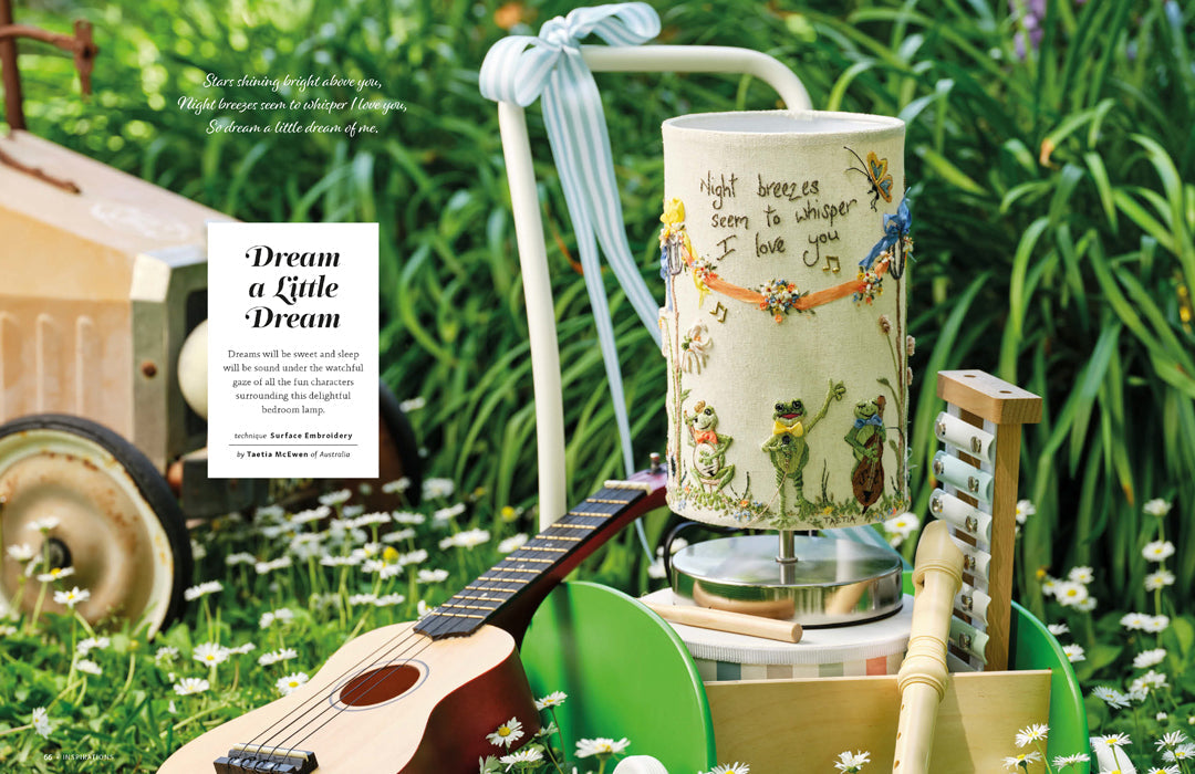 A decorative table lamp with a cream-colored drum shade featuring whimsical illustrations of children, bees, and handwritten text, styled in an outdoor garden setting next to a guitar and a book titled 'Dream a Little Dream'.
Photo by InspirationsStudios.com [additional_image_link]