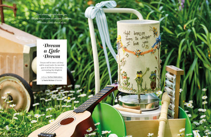 A decorative table lamp with a cream-colored drum shade featuring whimsical illustrations of children, bees, and handwritten text, styled in an outdoor garden setting next to a guitar and a book titled 'Dream a Little Dream'.
Photo by InspirationsStudios.com [additional_image_link]