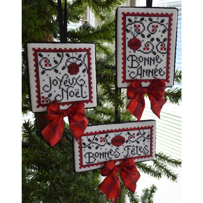 Three square cross-stitch Christmas ornaments hanging on a green Christmas tree. The ornaments feature festive French greetings ('Joyeux Noel', 'Bonne Annee', 'Bonnes Fetes') in black thread, decorated with red and black floral patterns, and tied with red satin bows.
Photo by Handsondesign.com [image_link]