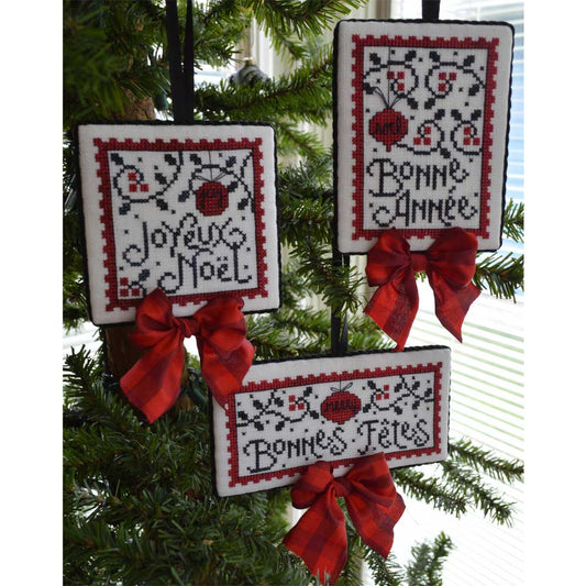 Three square cross-stitch Christmas ornaments hanging on a green Christmas tree. The ornaments feature festive French greetings ('Joyeux Noel', 'Bonne Annee', 'Bonnes Tentes') in black thread, decorated with red and black floral patterns, and tied with red satin bows.
Photo by Handsondesign.com [image_link]