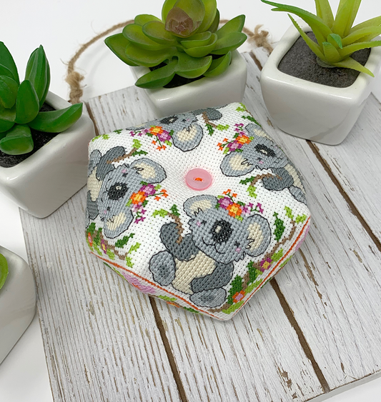 A small hexagonal cross-stitch cushion featuring a cute koala bear and floral pattern on a white fabric base, styled on a rustic white wooden surface with potted succulents in the background.
Photo by Tiny Modernist [image_link]