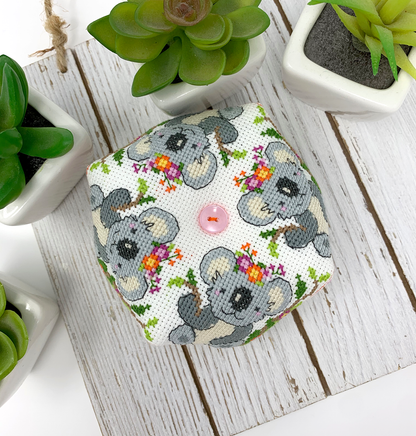 A small square fabric-covered item, likely a pouch or organizer, featuring a printed pattern of gray koalas with colorful floral crowns on a white background. The item has a small pink button on top and is photographed from above on a white wooden surface, surrounded by potted green succulents.
Photo by Tiny Modernist [additional_image_link]