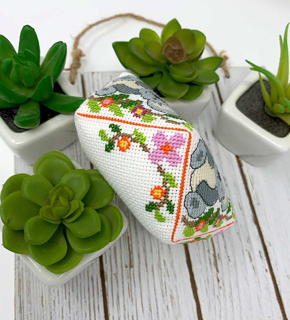 A small rectangular cross-stitch cushion with a white base, colorful floral vine embroidery, and gray animal motifs, styled on a white wooden surface among small potted succulents.
Photo by Tiny Modernist [additional_image_link]