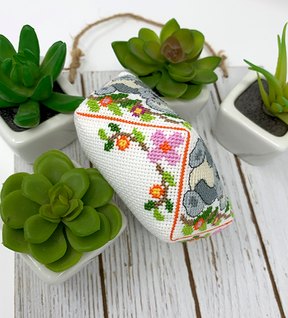 A small rectangular cross-stitch cushion with a white base, colorful floral vine embroidery, and gray animal motifs, styled on a white wooden surface among small potted succulents.
Photo by Tiny Modernist [additional_image_link]