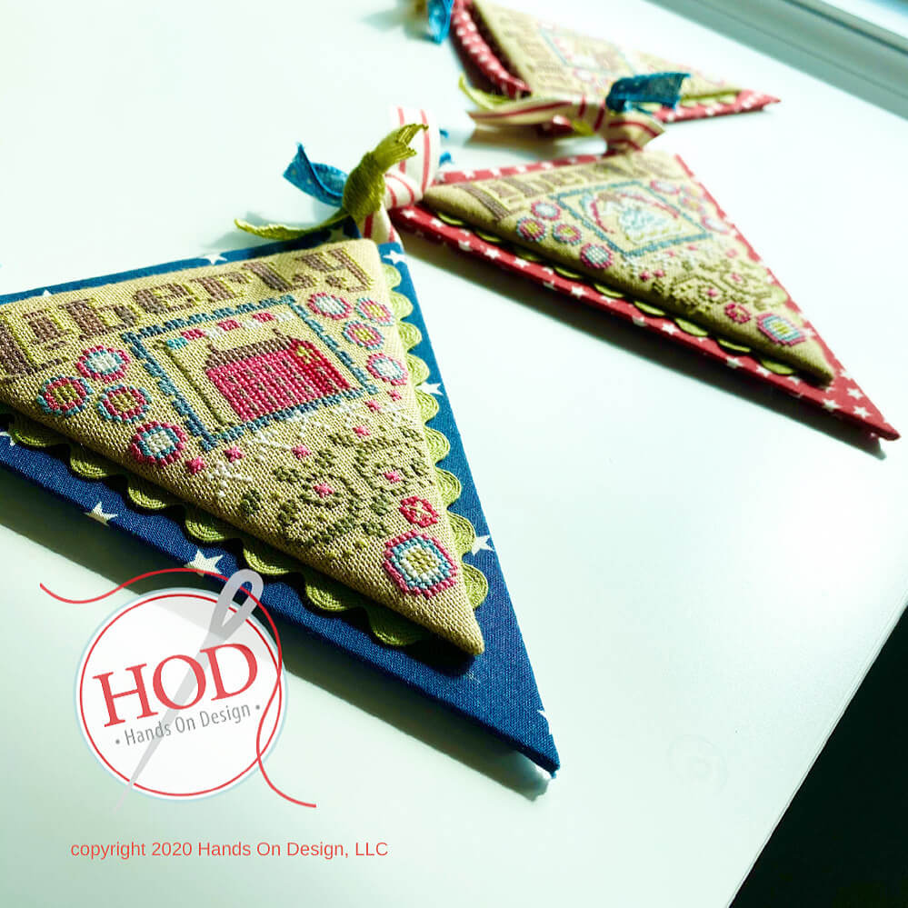 Two triangular embroidered fabric pieces displayed on a white surface, featuring detailed multicolor cross-stitch patterns on tan and red fabrics with blue ribbon ties. 'HOD Hands On Design'.
[additional_image_link]