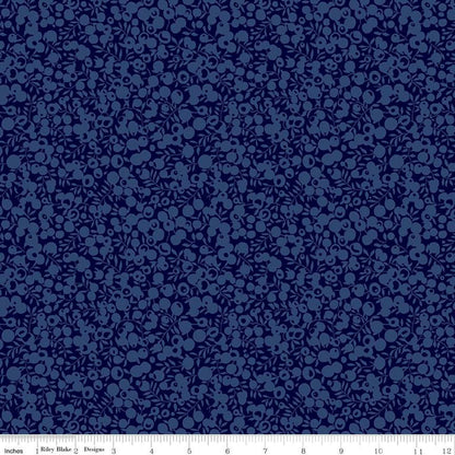 A close-up of a navy blue fabric with a repeating small floral or berry-like pattern in a slightly lighter blue. A white ruler strip is visible along the bottom edge with inch markings and the text 'Riley Blake Designs'.
[additional_image_link]