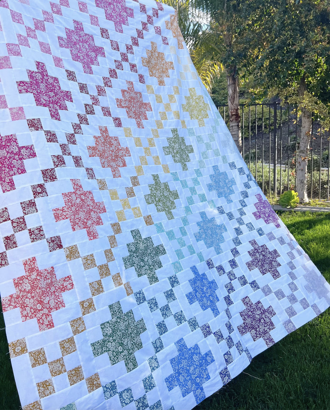 A large quilted blanket or throw laid outdoors on grass, featuring a white base with repeating geometric star-like motifs in pink, orange, yellow, green, blue, and purple, plus small checkered accents. Trees and a fence are visible in the background.
Photo by Jedi Craft Girl [image_link]