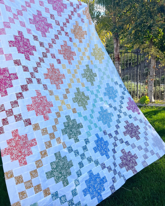 A large quilted blanket or throw laid outdoors on grass, featuring a white base with repeating geometric star-like motifs in pink, orange, yellow, green, blue, and purple, plus small checkered accents. Trees and a fence are visible in the background.
Photo by Jedi Craft Girl [image_link]