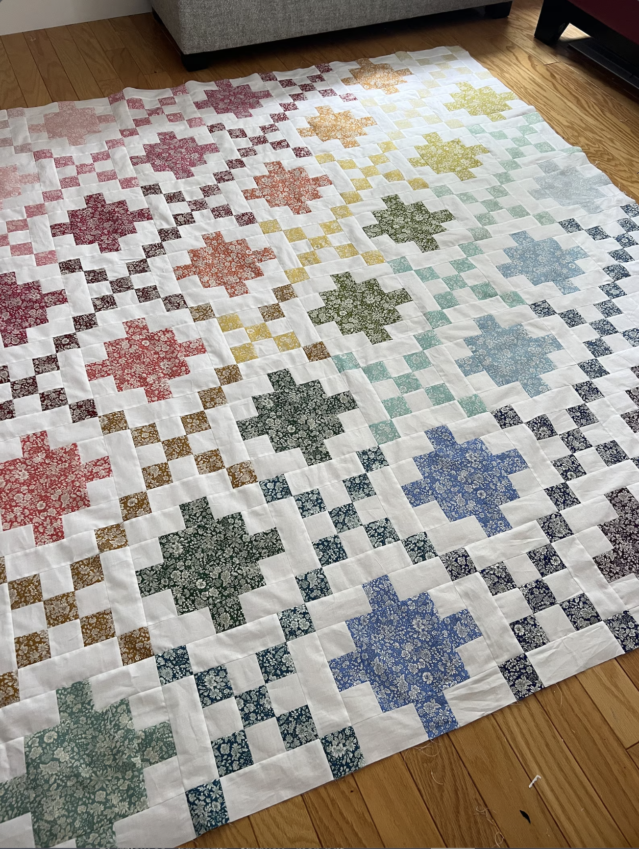 A handmade quilt laid out on a wooden floor, featuring a white base with a gradient of multicolor floral starburst blocks transitioning from pink and red on the left to yellow, green, and blue on the right, bordered by small patterned checkered squares.
Photo by Jedi Craft Girl [additional_image_link]