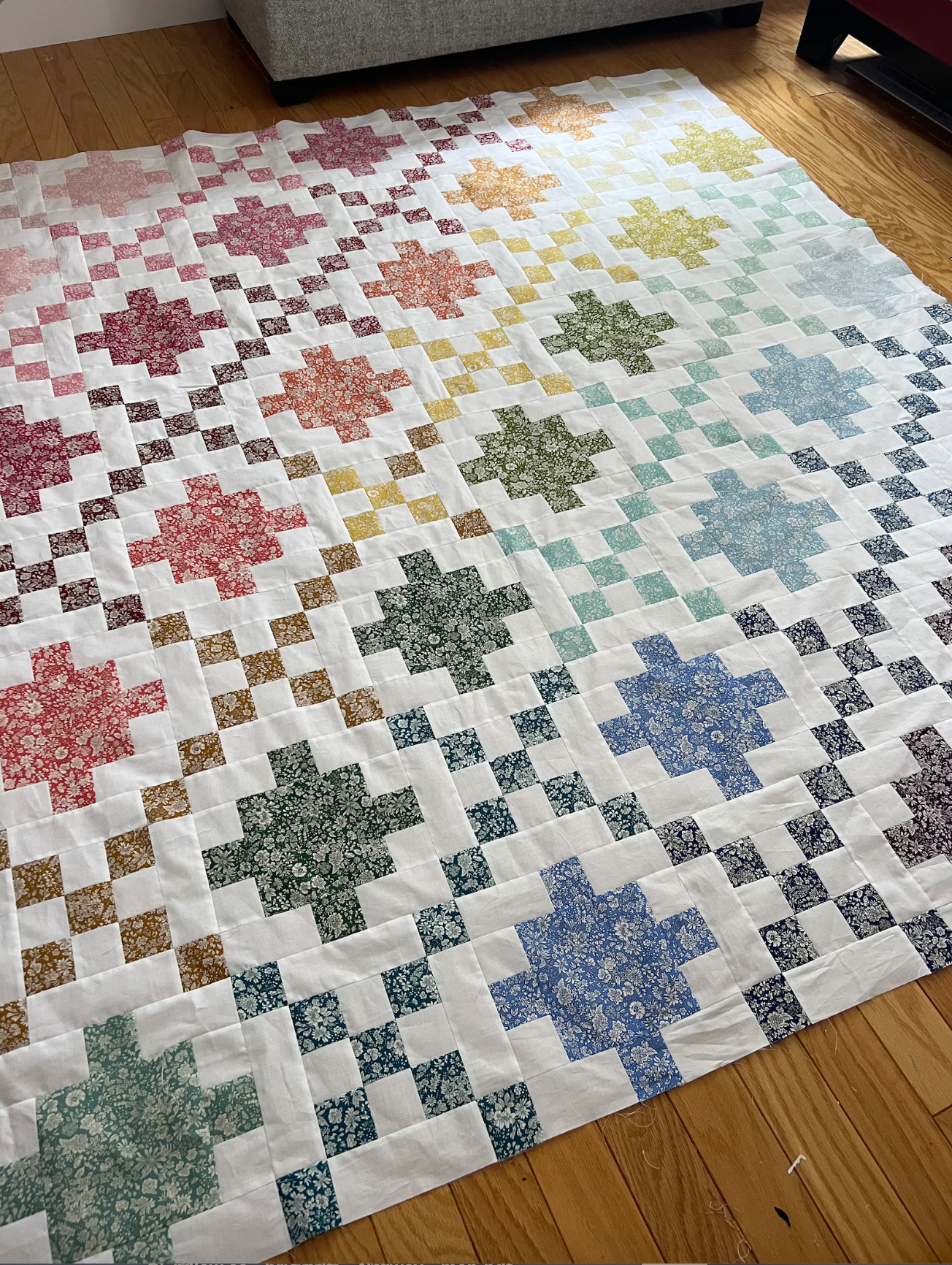 A handmade quilt laid out on a wooden floor, featuring a white base with a gradient of multicolor floral starburst blocks transitioning from pink and red on the left to yellow, green, and blue on the right, bordered by small patterned checkered squares.
Photo by Jedi Craft Girl [additional_image_link]