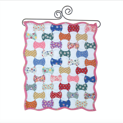 A quilted wall hanging featuring a pattern of red and green applecores on a white background, with a pink border at the top.
Photo by Sue Daley [image_link]