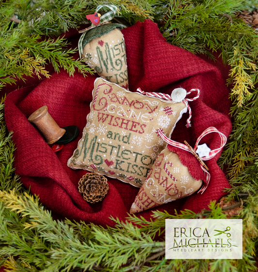 Three embroidered Christmas ornament pillows arranged on a red knit blanket surrounded by pine branches. The central pillow reads 'Candy Cane Wishes and Mistletoe Kisses' with red embroidery and white stitching.
Photos by Erica Michaels [image_link]