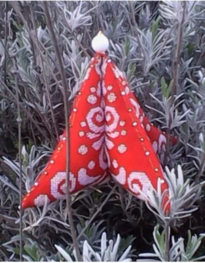 A red and white cross stitch ornament with a Christmas tree design, made with Jobelan fabric and adorned with beads, resting on a plant with green foliage in the background.
[image_link]