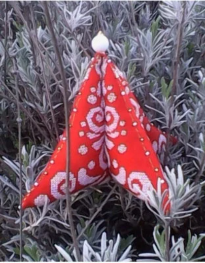 A red and white cross stitch ornament with a Christmas tree design, made with Jobelan fabric and adorned with beads, resting on a plant with green foliage in the background.
[image_link]