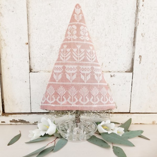 A small decorative needlepoint Christmas tree with a dusty pink and white Nordic pattern, displayed on a clear glass base with white flowers and green leaves in front of a rustic white wooden background.
Photo by HelloFromLizMathews.com [image_link]