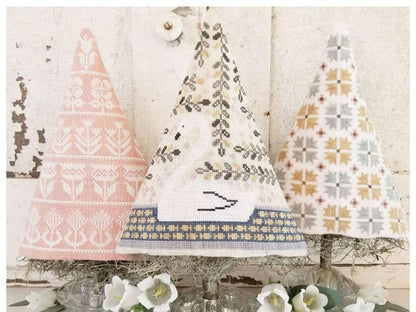 Three decorative needlepoint Christmas tree ornaments in pink, white, and beige patterns, displayed on a rustic white wooden background with white flowers at the bottom.
Photo by HelloFromLizMathews.com [additional_image_link]