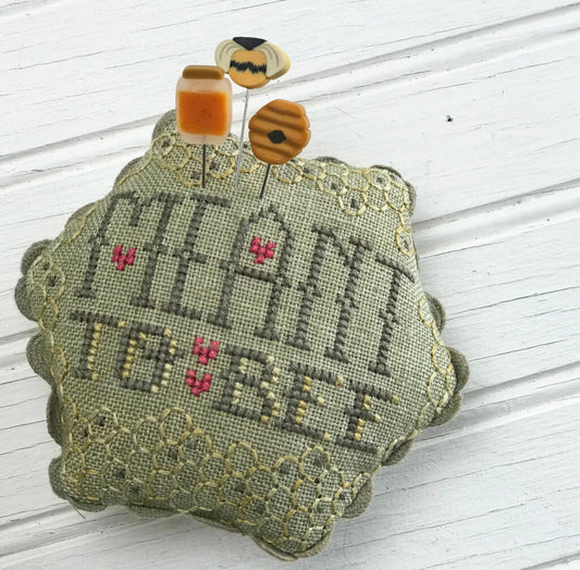 A decorative cross-stitch cushion with the words 'MEANT TO BE' stitched in gray thread, accented with small pink flowers and a scalloped olive-green lace border. Two small bees on pins rest on top, set against a white wooden background.
Photo by Hands on Design [image_link]
