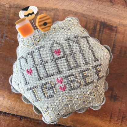 A handmade cross-stitch sampler pillow on a rustic wooden background. The pillow is made of light beige linen fabric with scalloped edges and features the words 'MEANT TO BE' stitched in gray thread, accented with small pink hearts.
Photo by Hands on  Design [additional_image_link]
