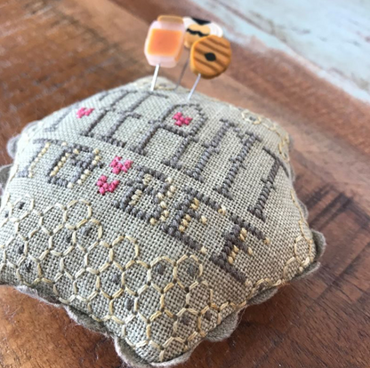 A small embroidered cross-stitch pillow on a rustic wooden surface, featuring a bee and floral design in muted gray, beige, and pink thread.
Photo by Hands on  Design [additional_image_link]